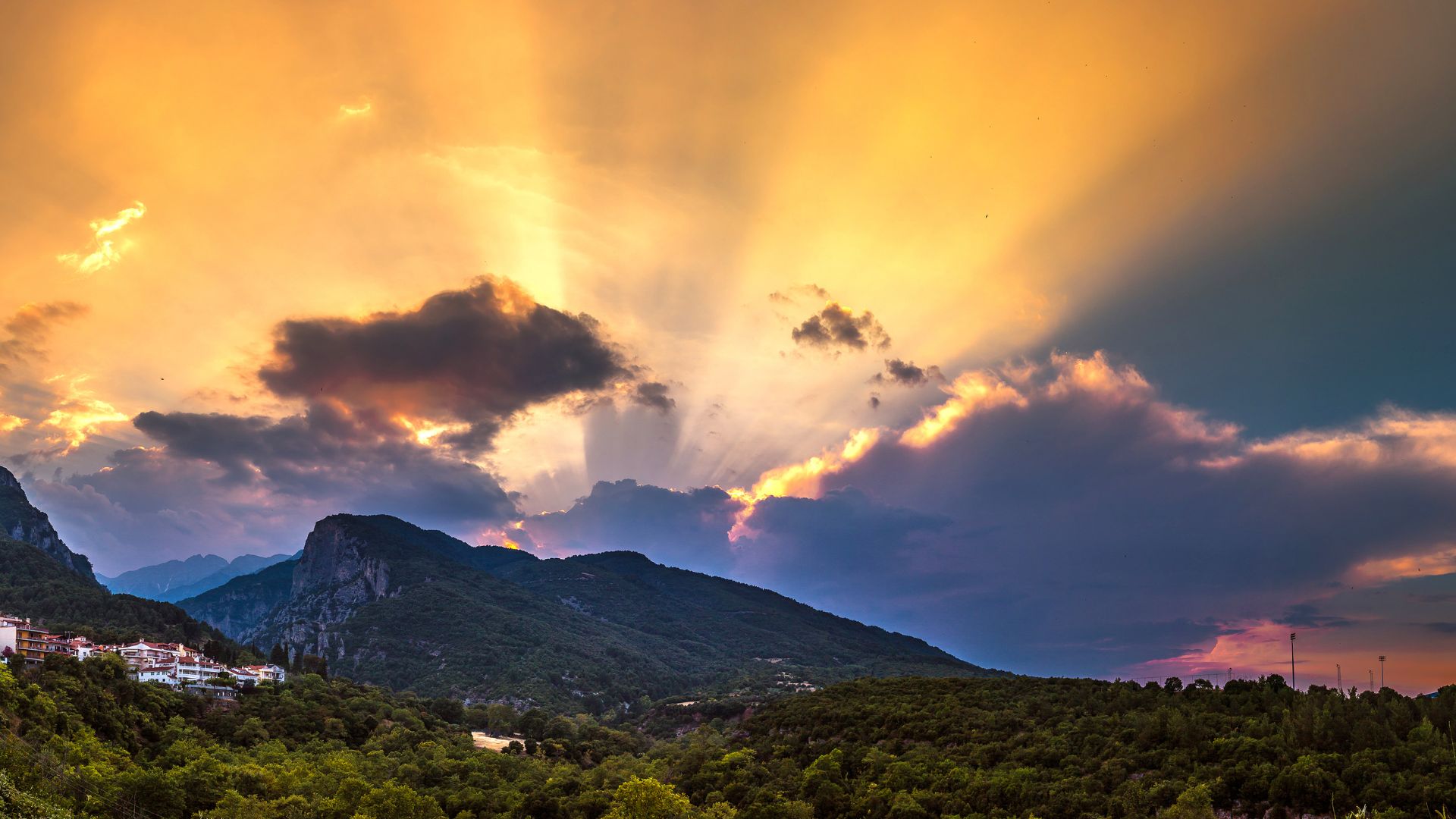 A dramatic sunset over Mount Olympus, Greece, with vibrant orange and purple clouds illuminated by sunbeams piercing through the sky, casting light over the lush green mountainside and a small village nestled in the foothills.