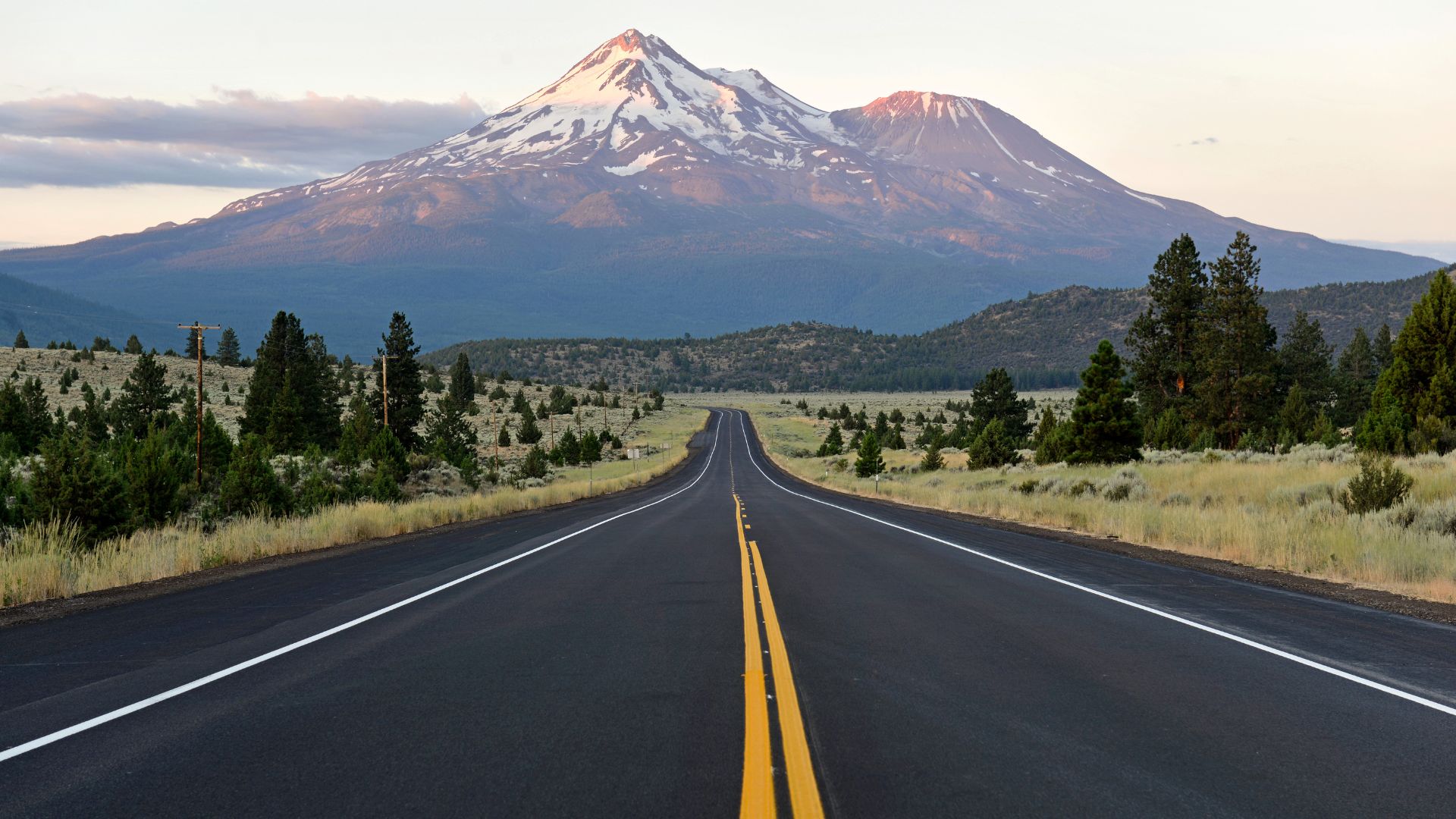 A long, straight road leads towards a majestic, snow-capped Mount Shasta, a prominent stratovolcano in Northern California, under a partially cloudy sky. The foreground features dry grass and scattered pine trees on either side of the road.