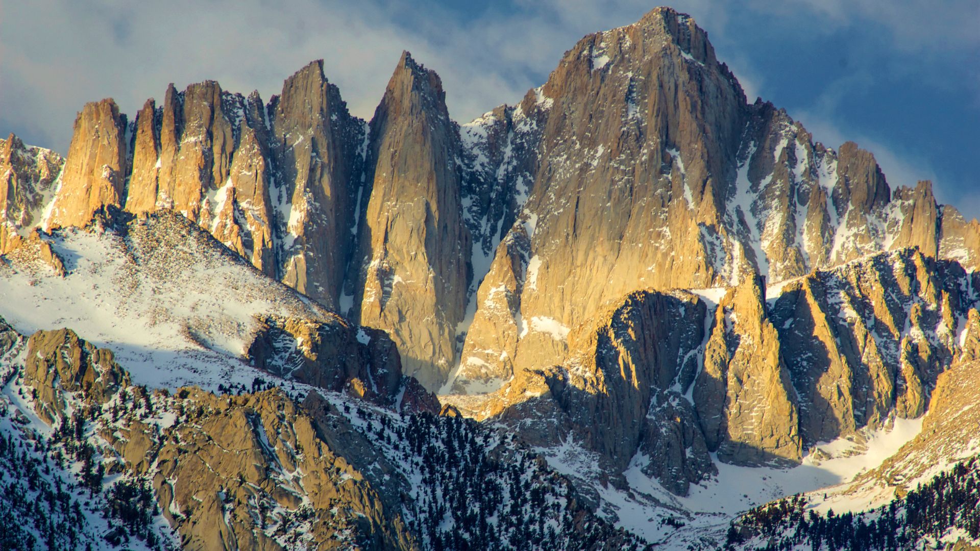 A majestic panoramic view of Mount Whitney, the highest peak in the contiguous United States, showcasing its rugged, snow-dusted granite peaks under a dramatic sky with sunlight illuminating portions of the mountainside.