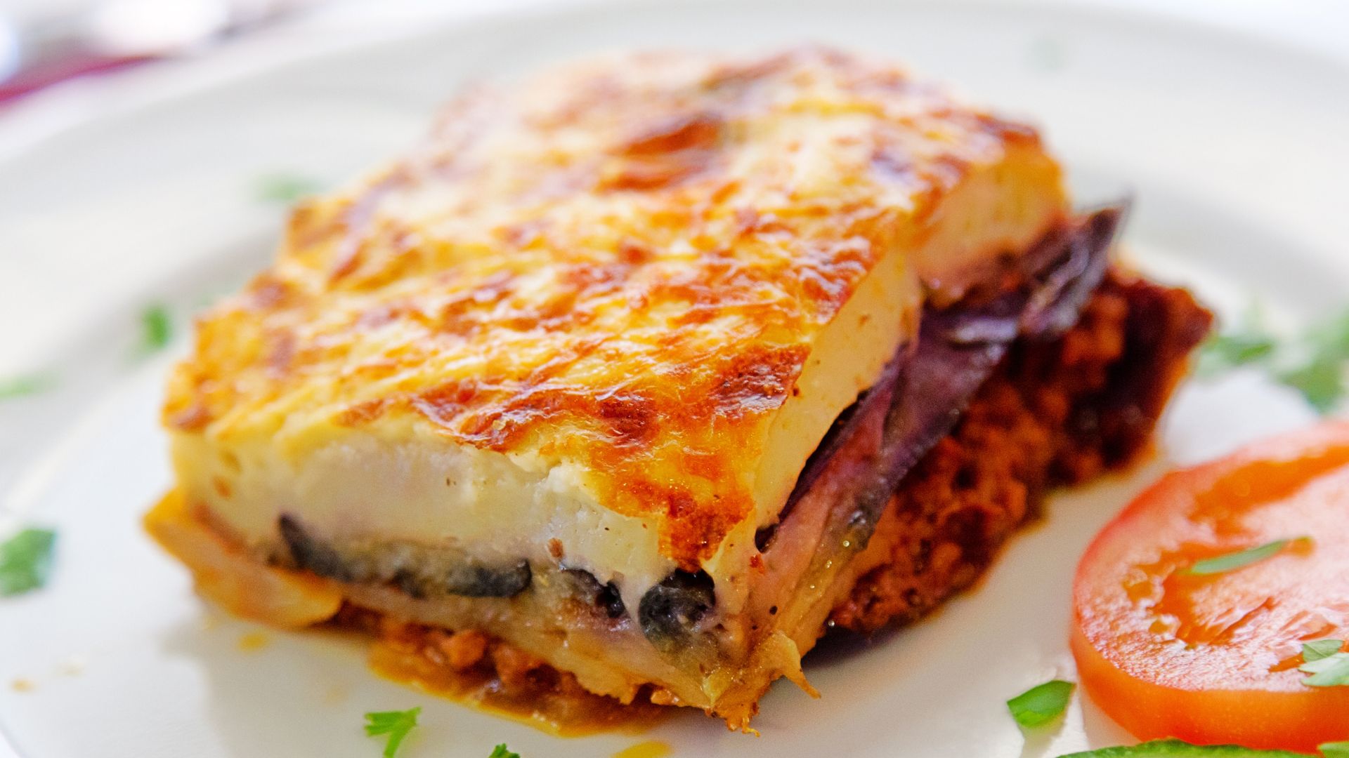 A close-up shot of a serving of moussaka, featuring layers of baked eggplant, meat sauce, and a creamy béchamel topping, garnished with a slice of tomato and parsley.