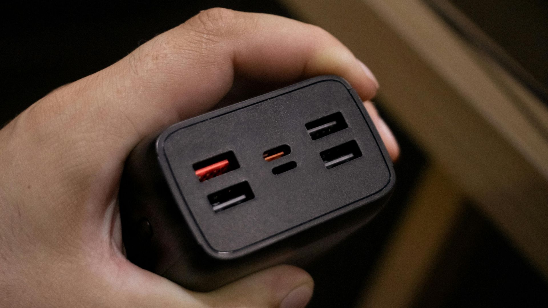 A hand holds a black power bank or multi-port charger, featuring several USB-A ports (two with red interiors, two black) and two smaller USB-C ports. 