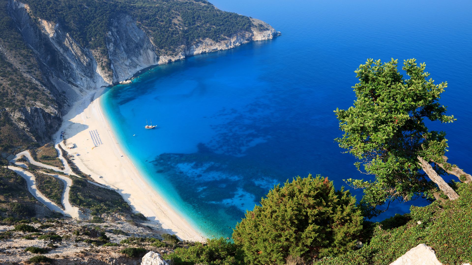 An aerial view of Myrtos Beach in Kefalonia, Greece, showcasing its crescent-shaped expanse of white pebbles, surrounded by dramatic cliffs, and meeting the vivid turquoise and deep blue waters of the Ionian Sea, with lush green trees in the foreground. 