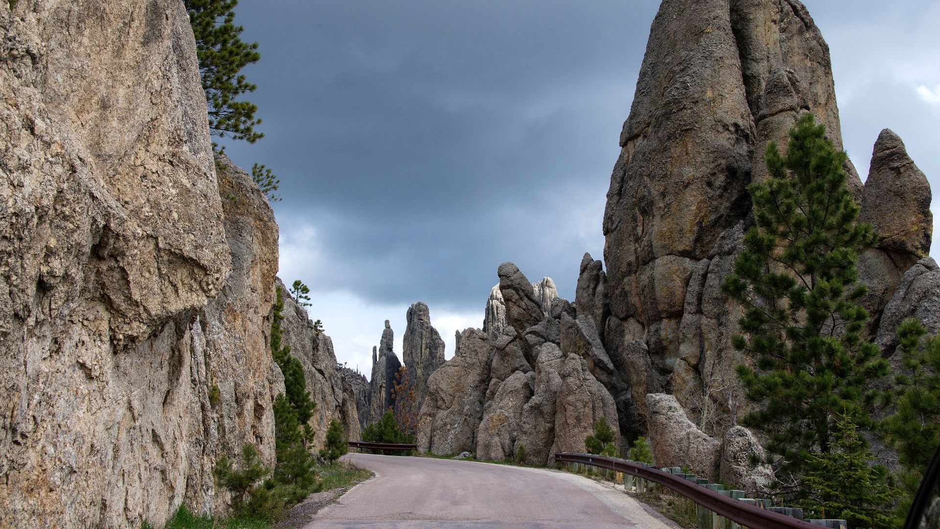A winding paved road, Needles Highway (South Dakota Highway 87), curves through a dramatic landscape of towering granite rock formations, often referred to as "needles," and scattered pine trees under a cloudy sky in Custer State Park.