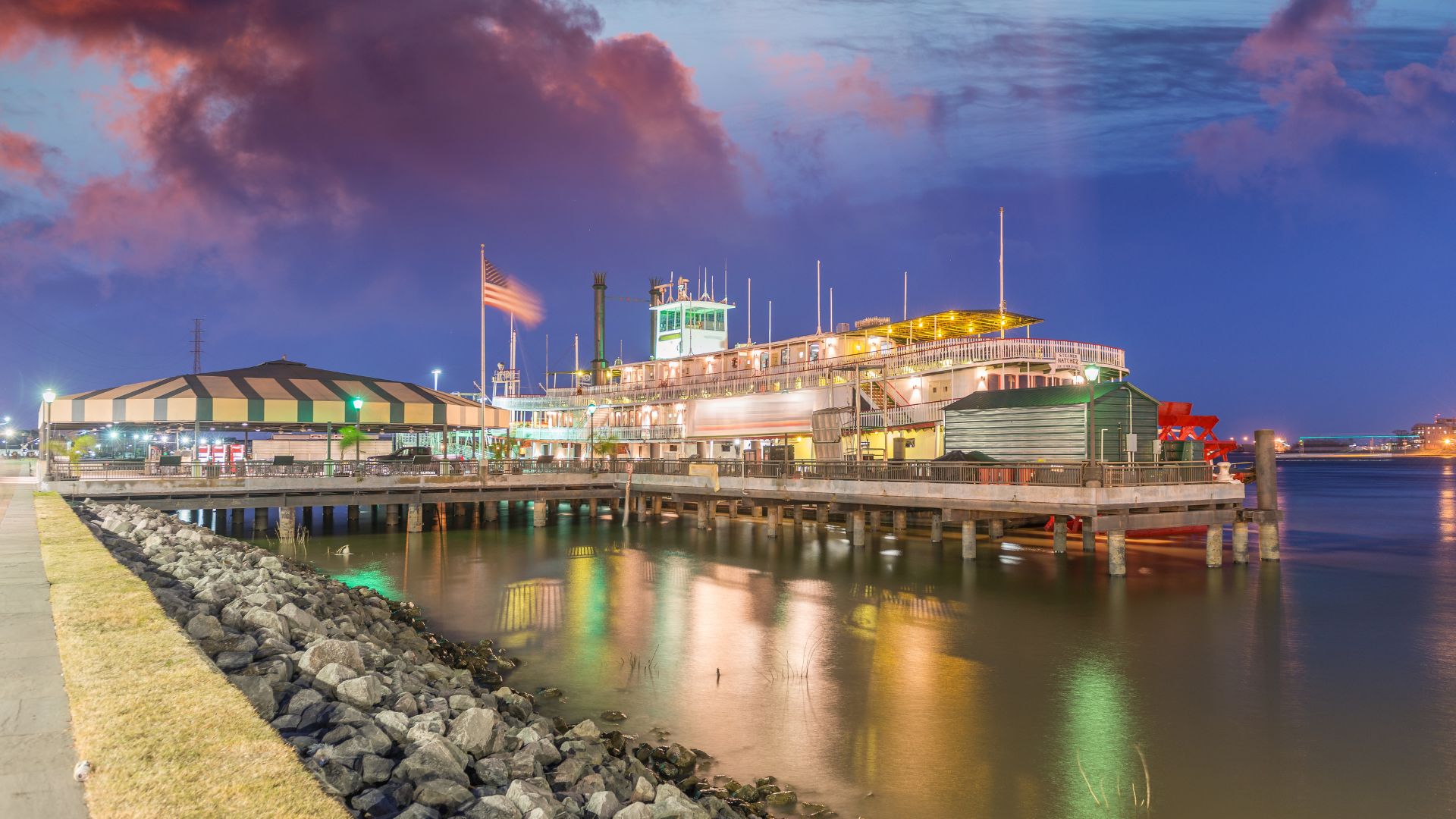 A wide shot at dusk shows the illuminated Steamboat Natchez, a large paddlewheel riverboat, docked at a pier on the Mississippi River in New Orleans, with a tented structure and an American flag visible on the pier, and a vibrant purple and pink sky above.