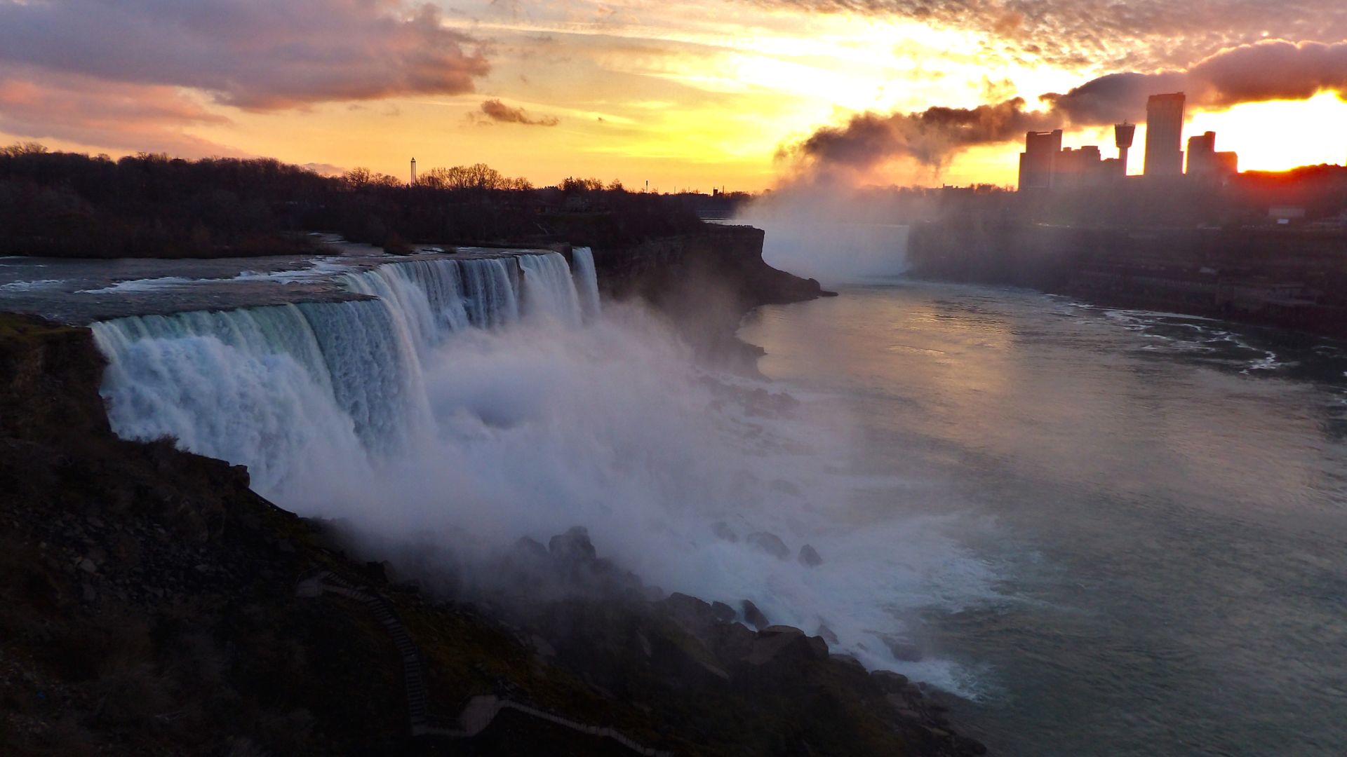 A wide-angle shot of Niagara Falls at sunset, showcasing the powerful waterfalls, the mist rising from the falls, and the Niagara River flowing towards a city skyline visible in the distance under a colorful, cloudy sky.