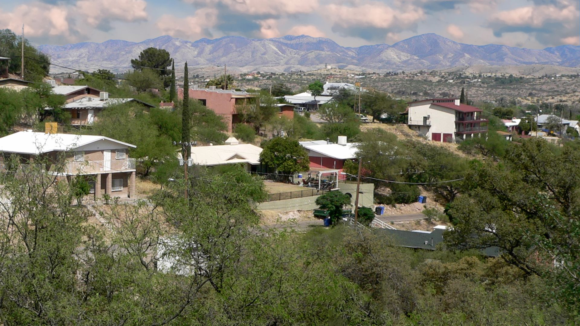 A panoramic view of a residential hillside in Nogales, Arizona, featuring houses nestled among green trees and shrubs, with a backdrop of distant mountains under a cloudy sky.