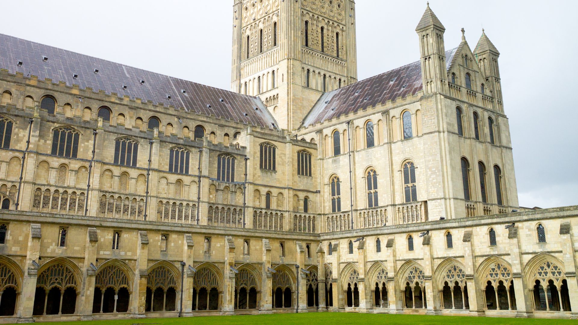 A wide-angle exterior view of Norwich Cathedral, showcasing its large, ornate stone structure and the expansive, well-maintained green lawn of the cloister in the foreground.