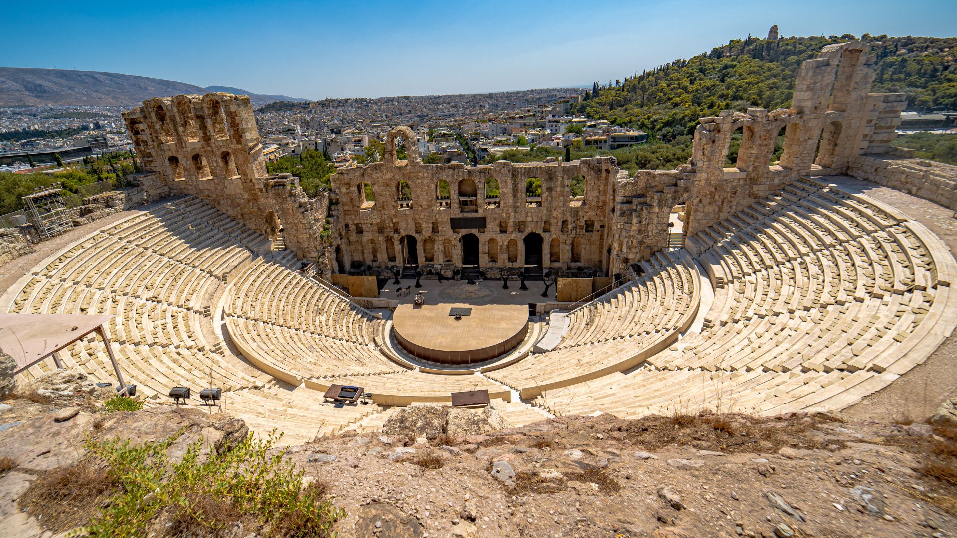 A wide shot of the ancient Odeon of Herodes Atticus, an amphitheater with tiered seating and a partially restored stage building, nestled at the base of the Acropolis hill in Athens, Greece, under a clear blue sky. 
