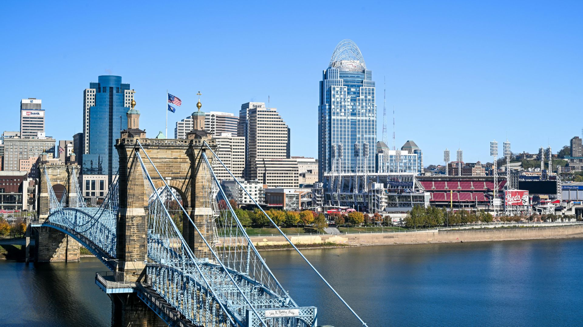 A panoramic view of the Cincinnati skyline with the iconic John A. Roebling Suspension Bridge in the foreground, stretching across the Ohio River, with various city buildings and a stadium visible on the riverbank under a clear blue sky.