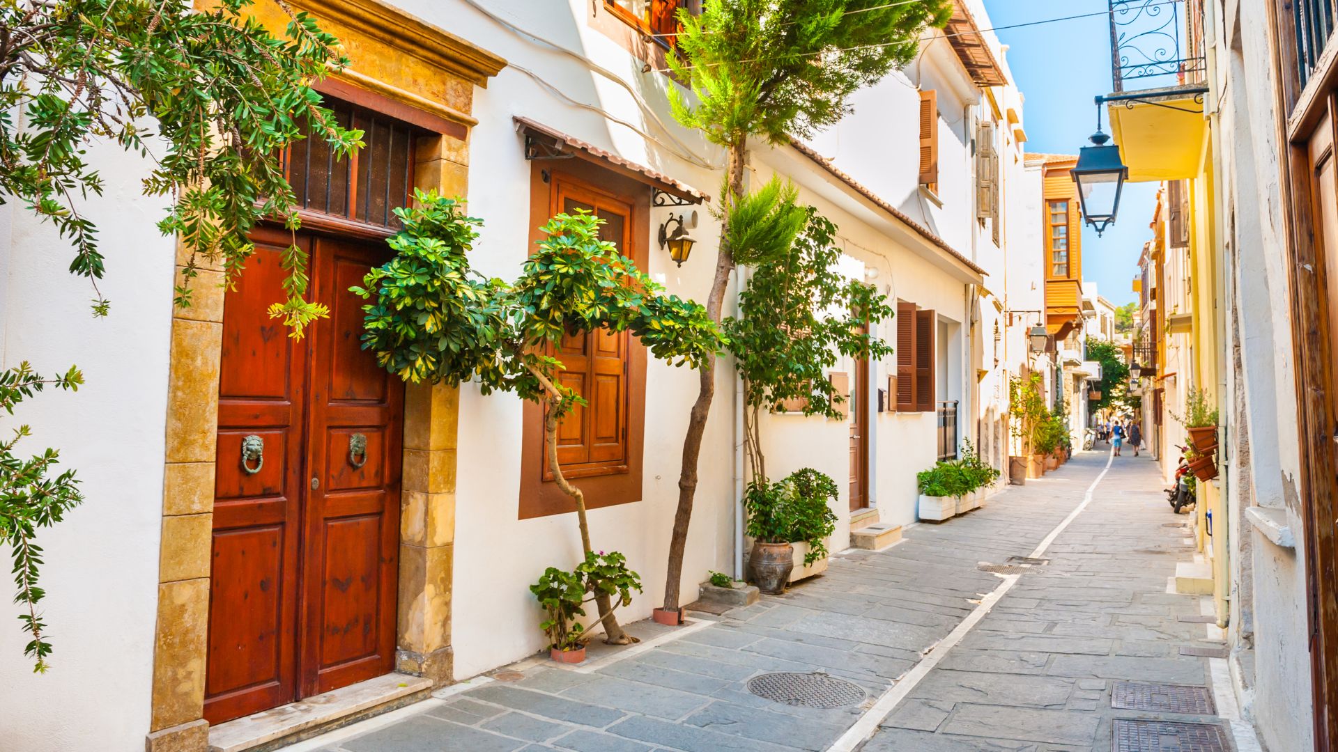 A narrow, cobbled street in Rethymno's Old Town, lined with white-walled buildings featuring traditional wooden doors and windows, potted plants, and small trees, under a clear blue sky.