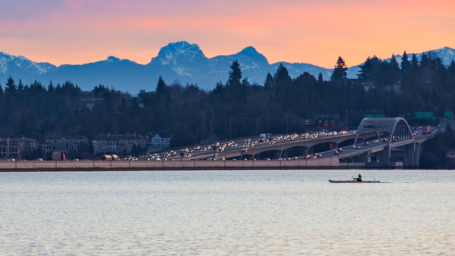 A wide shot of the I-90 floating bridge on Lake Washington at sunset, with mountains and forested hills in the background, and a lone kayaker on the water in the foreground. 