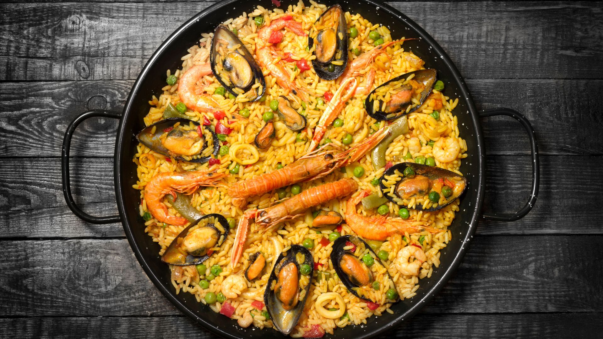 A close-up, overhead view of a large pan of seafood paella, richly colored with saffron rice, mussels, shrimp, and peas, ready to be served.