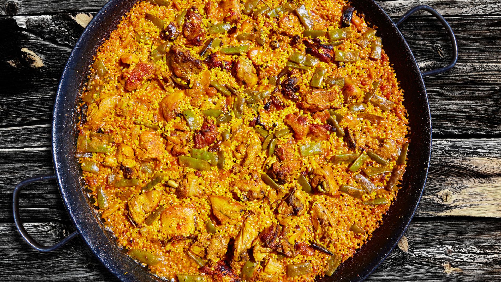 A close-up overhead shot of a large, shallow black paella pan filled with golden-orange paella, featuring chicken pieces, green beans, and rice, set against a rustic dark wooden background.