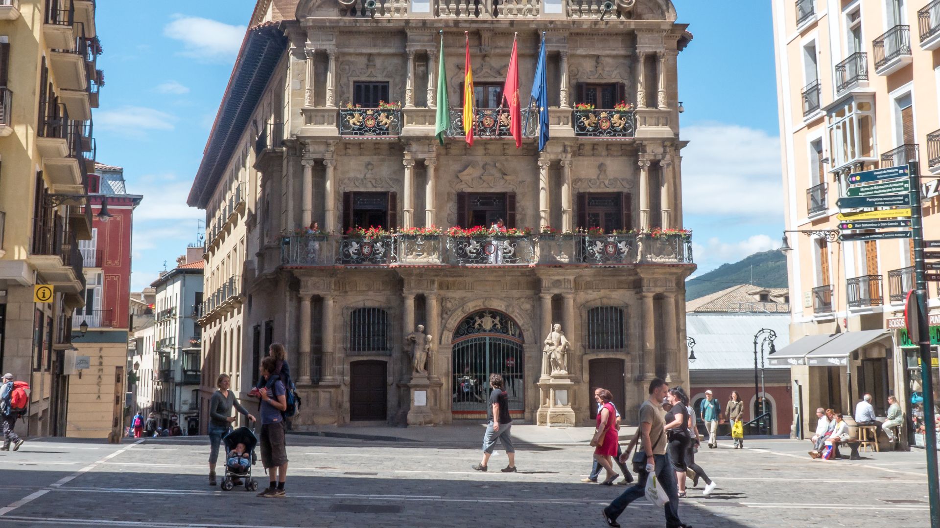 A wide-angle view of the ornate, multi-story Baroque facade of the Pamplona City Hall, with flags hanging from the balconies, situated in a bustling city square with people walking and gathered in groups.
