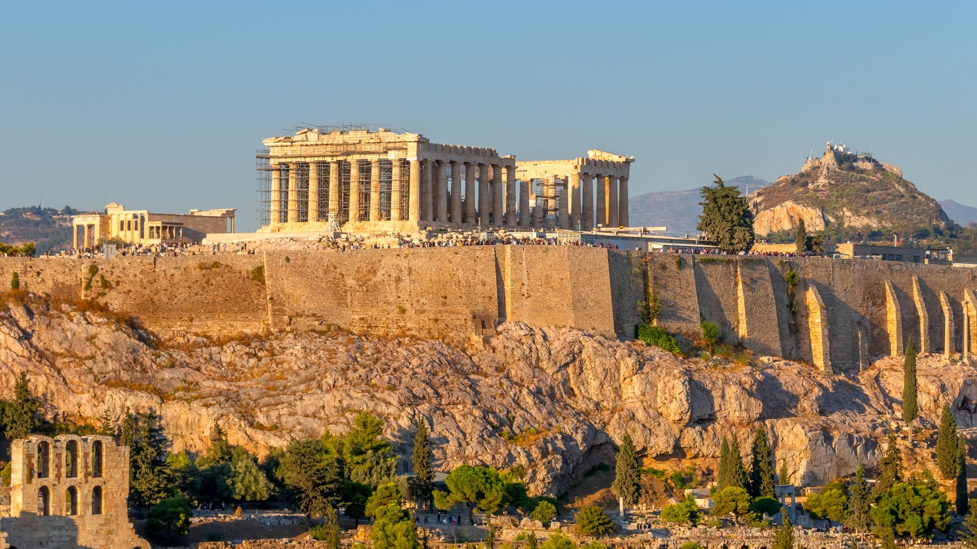 A wide-angle view of the ancient Parthenon temple atop the rocky Acropolis hill in Athens, Greece, with the city sprawling below and distant mountains under a soft, clear sky.
