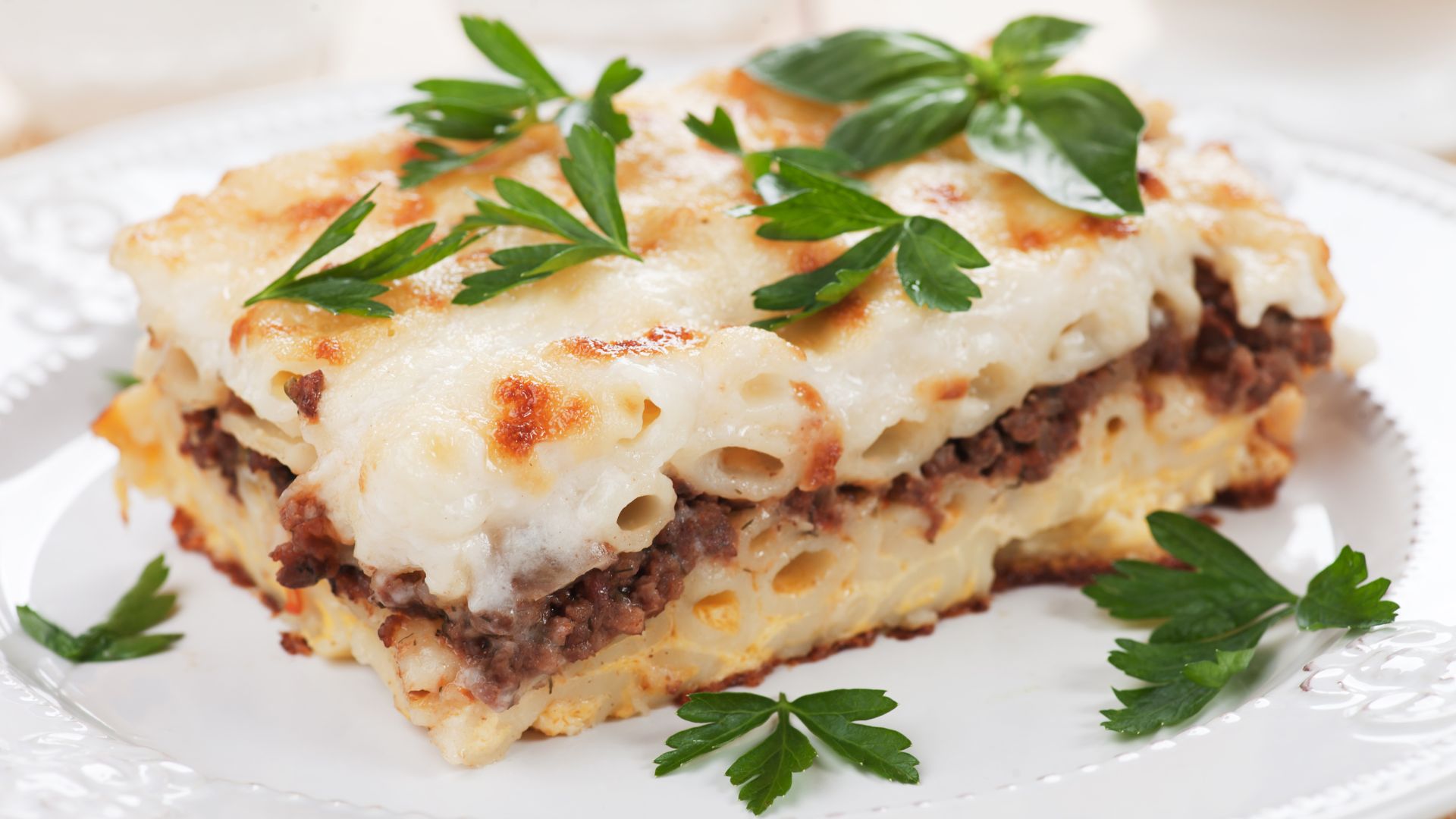 A close-up of a serving of baked Greek Pastitsio, featuring layers of macaroni pasta, seasoned ground meat, and a creamy béchamel sauce, topped with melted cheese and garnished with fresh parsley. 