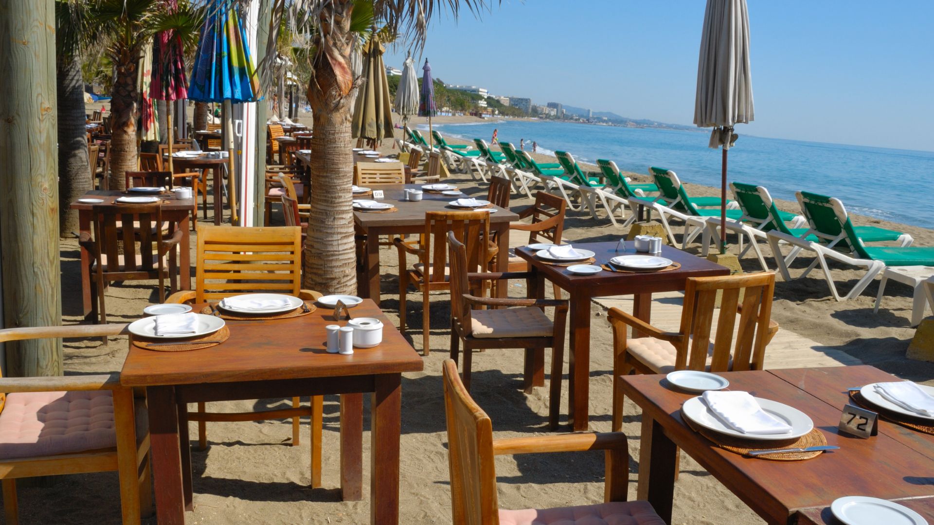 A beachfront restaurant scene with wooden tables and chairs set up on the sand, facing the calm blue sea. Palm trees and beach umbrellas provide shade, and sun loungers are visible along the shoreline in the background.