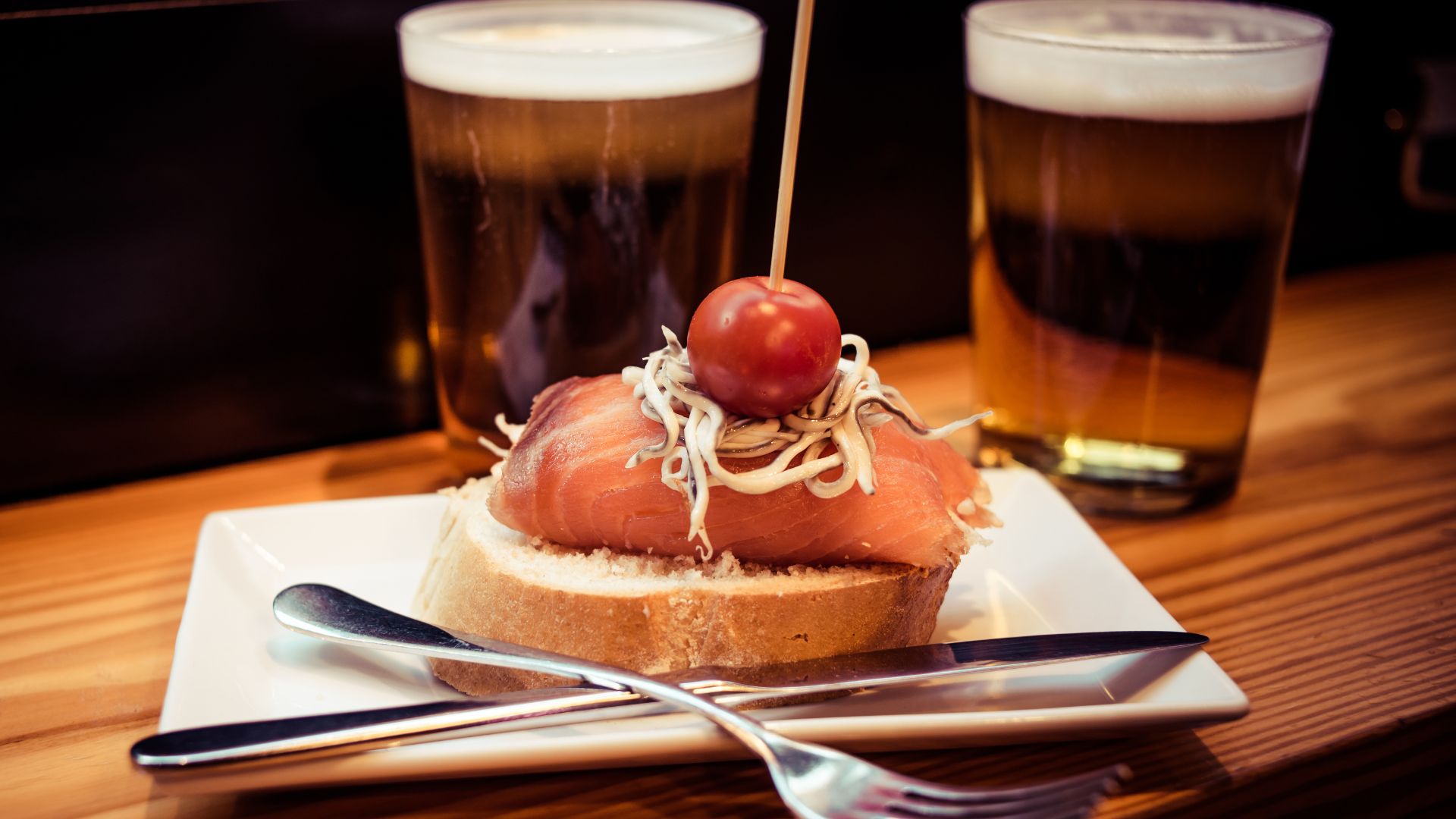 A close-up shot of a pintxo, a small snack consisting of a piece of bread topped with cured meat, shredded ingredients, and a cherry tomato held together by a toothpick, with blurred glasses in the background.