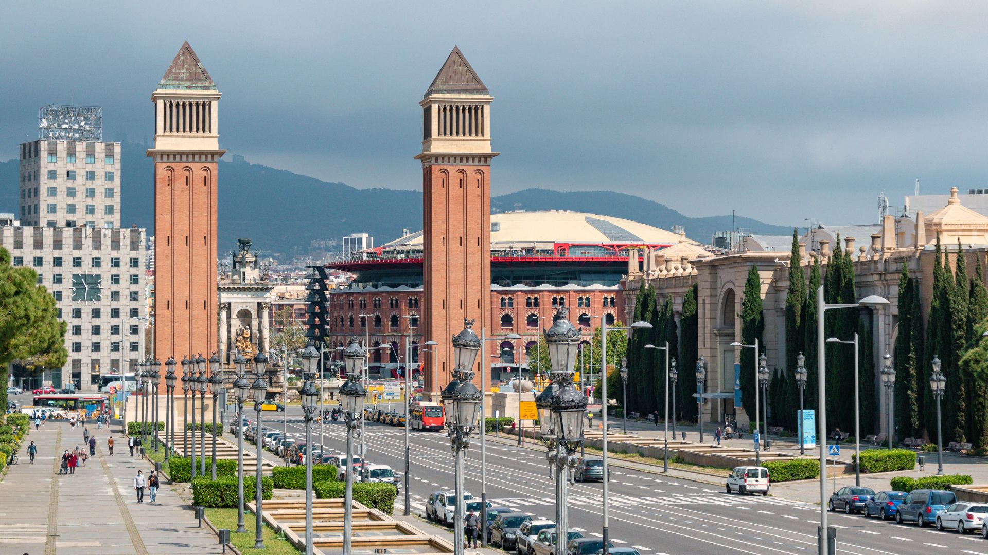 The image depicts the Plaça d'Espanya in Barcelona, featuring the prominent Venetian Towers flanking the Avinguda de la Reina Maria Cristina, with the former bullring Las Arenas (now a shopping center) visible in the background, all set against a backdrop of distant hills and a clear sky. 