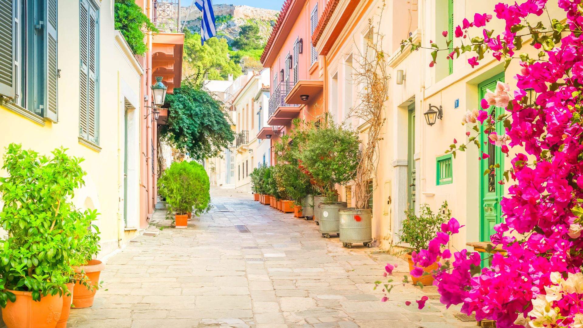 A narrow, cobblestone street in Athens' Plaka neighborhood, lined with colorful neoclassical buildings adorned with potted plants and vibrant bougainvillea, under a clear blue sky.