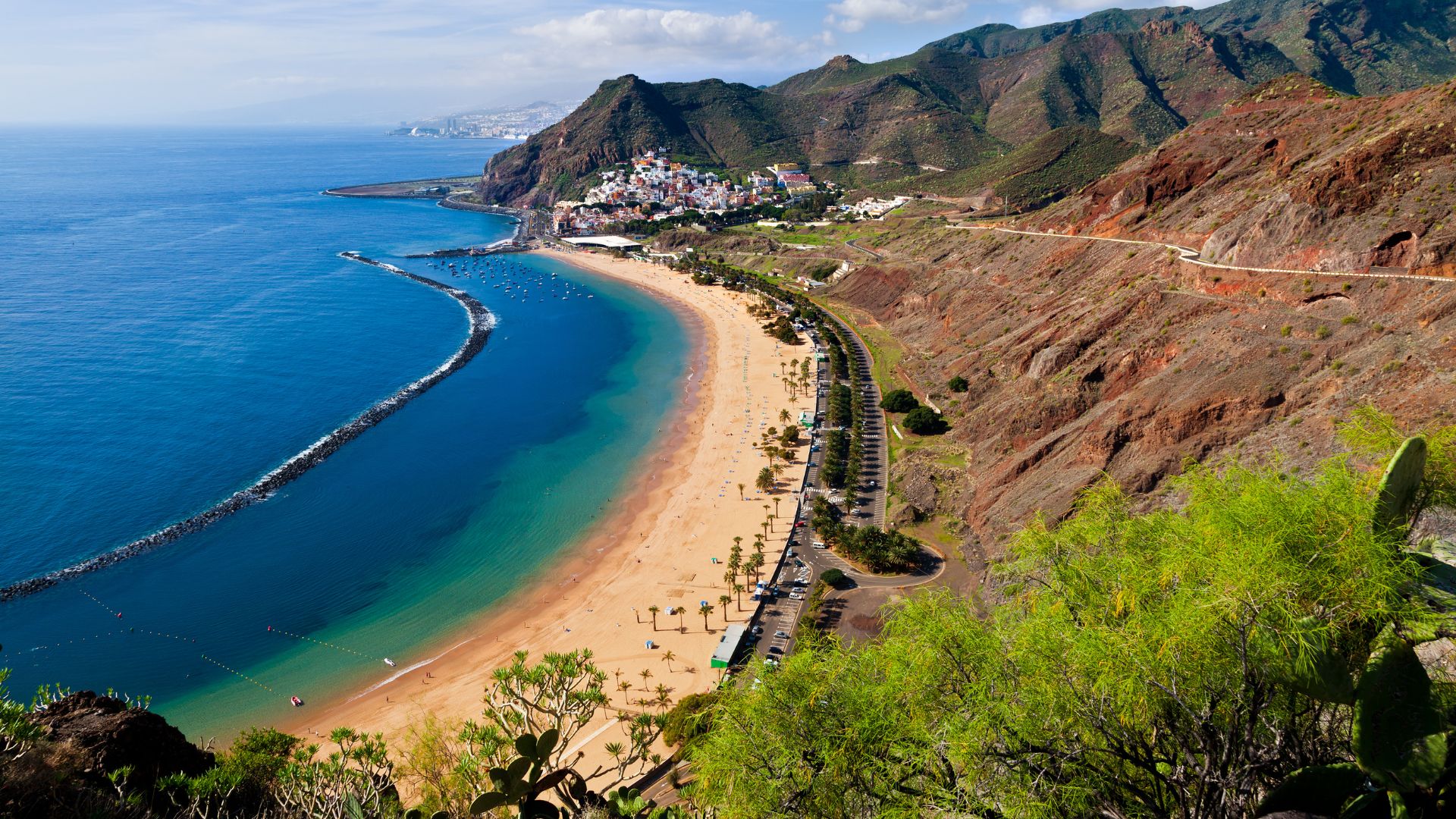 A wide, golden sand beach with calm turquoise waters, protected by a long breakwater, framed by lush green hills on one side and the deep blue ocean on the other, with a small town nestled at the base of the hills in the distance.
