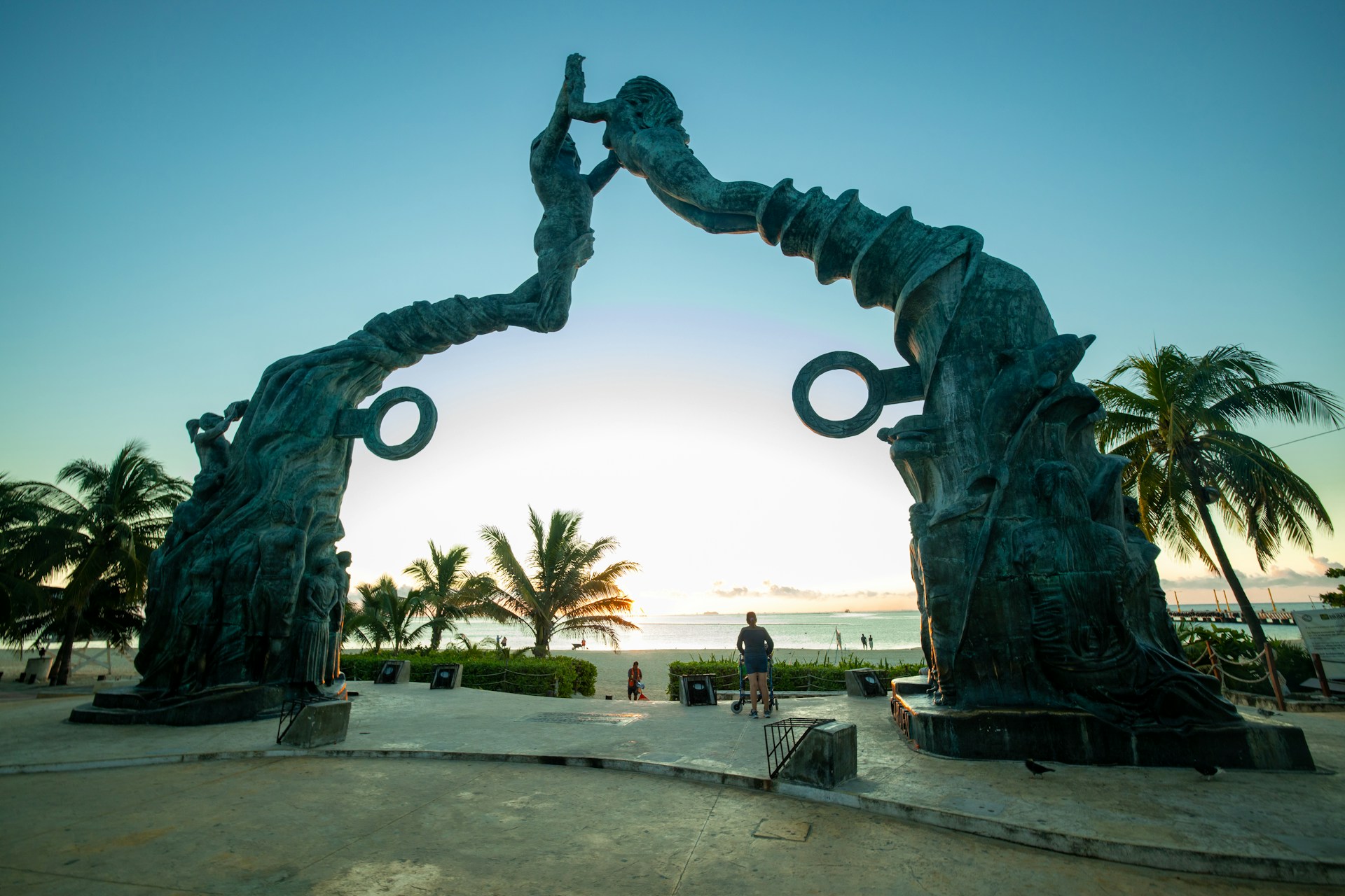 Large bronze sculpture in Playa del Carmen depicting a man and a woman with flowing, tree-like forms reaching upward to touch hands and form an arch, set against the Caribbean Sea at Parque Los Fundadores.