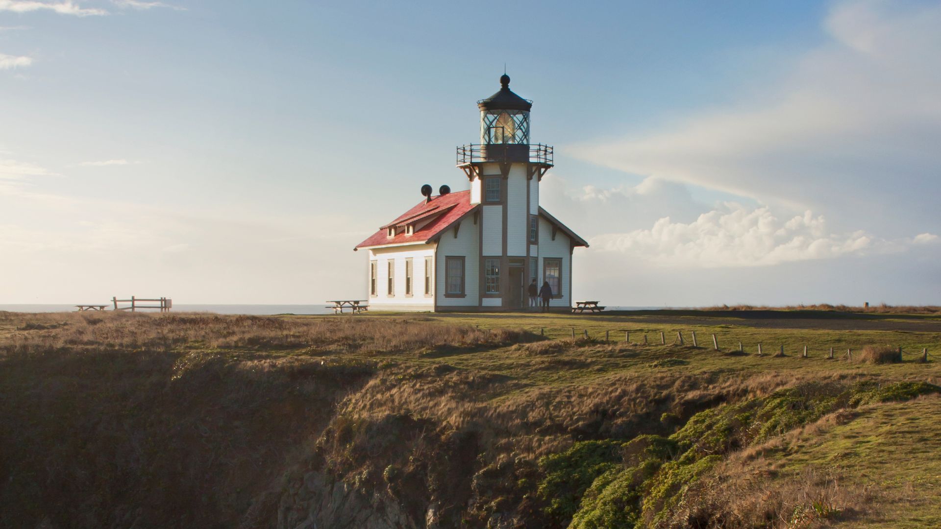 A historic white lighthouse with a dark roof and a prominent lantern room stands on a grassy headland under a bright, sunlit sky, with the ocean visible in the background.