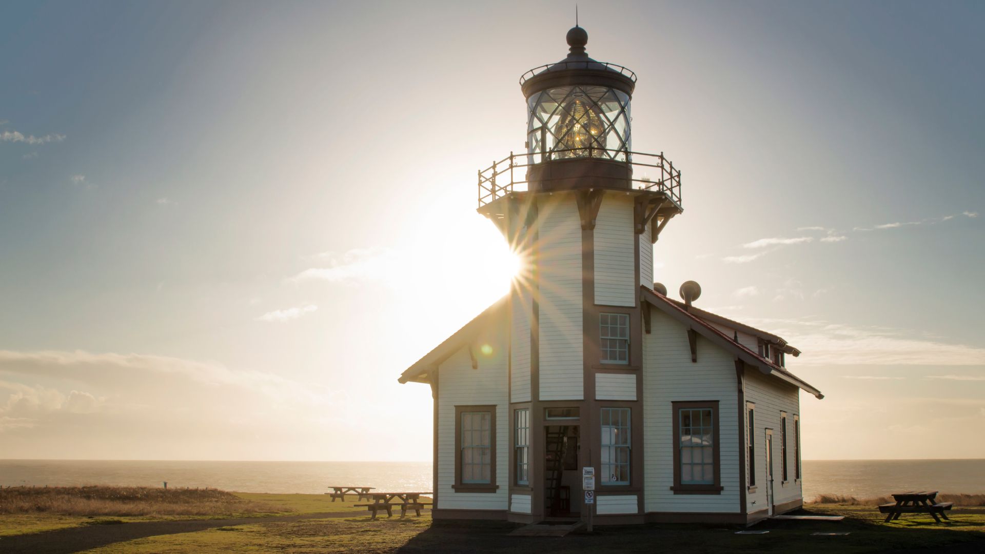 A historic white lighthouse with a dark roof and a prominent lantern room stands on a grassy headland under a bright, sunlit sky, with the ocean visible in the background.