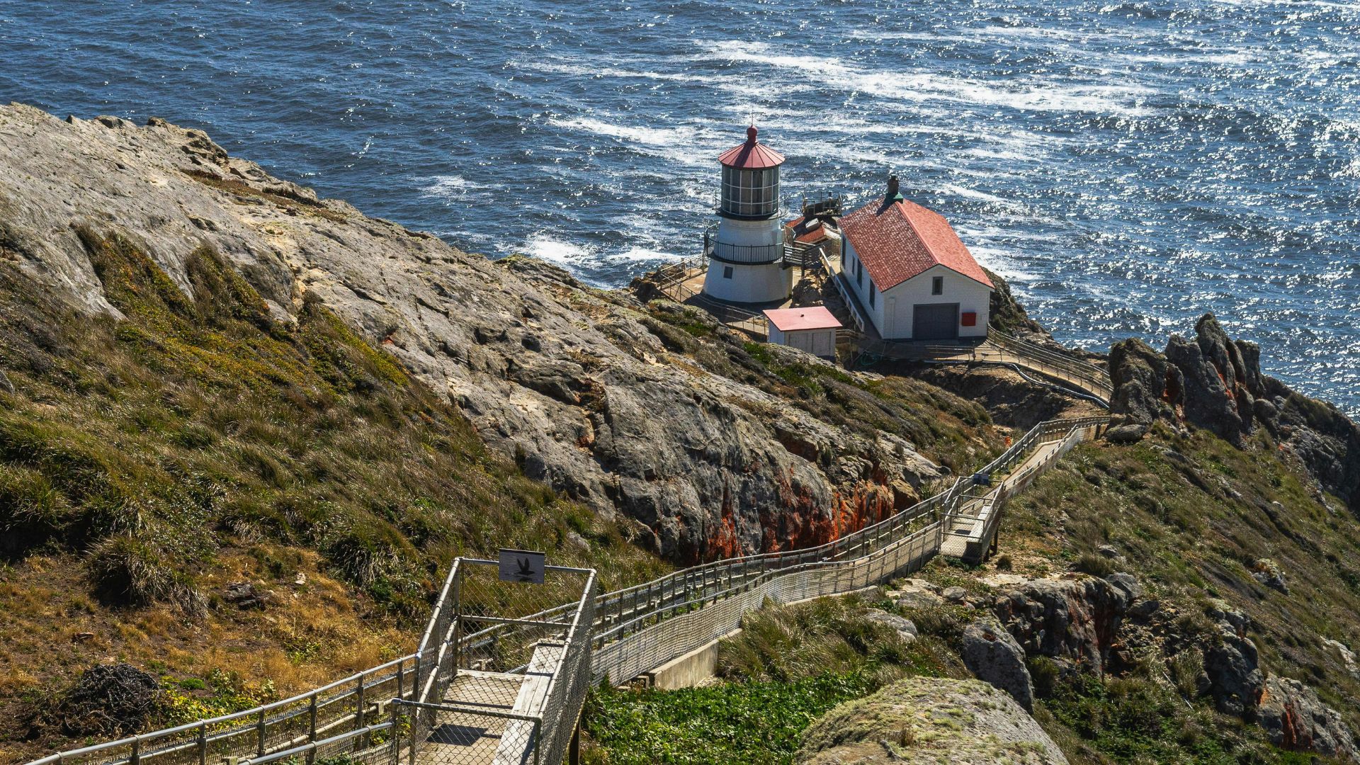 A dramatic aerial view of the Point Reyes Lighthouse and its adjacent buildings perched on a rugged cliffside, connected by a long, winding staircase and pathway leading down to the ocean.