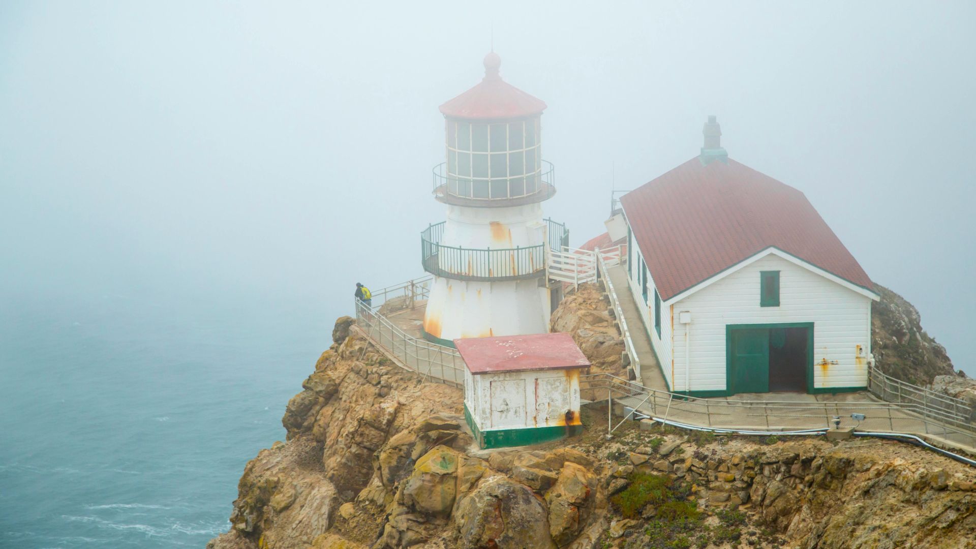 A lighthouse with a red roof and an adjacent white building perched on a rocky cliff overlooking the ocean, shrouded in fog.