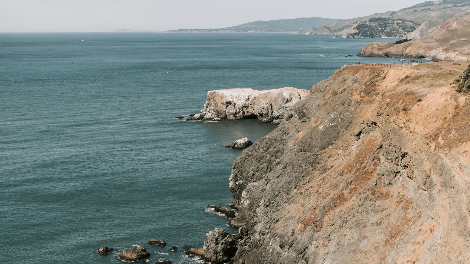 A high-angle view of a secluded beach at the base of steep, rocky cliffs, with waves breaking on the dark sand and the vast ocean stretching into the distance.