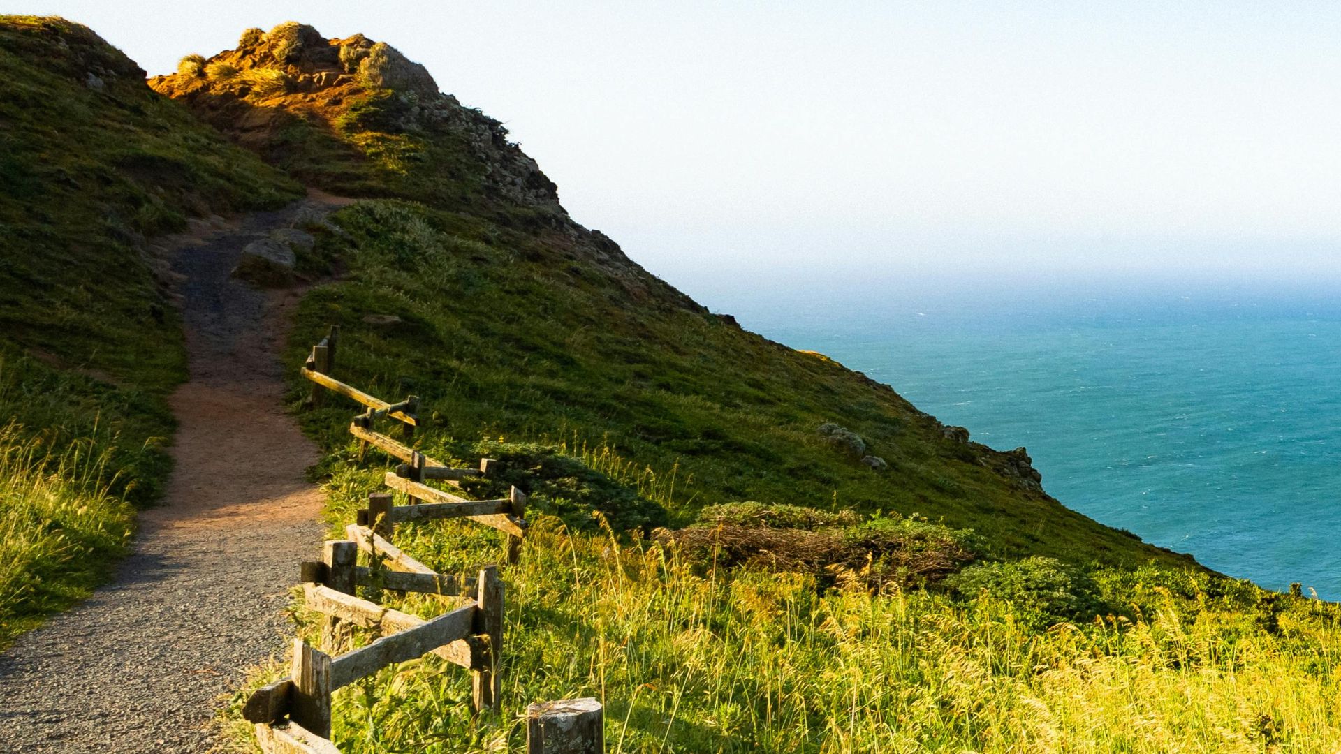 A winding dirt path with a rustic wooden fence on the left, leading up a green, grassy hillside overlooking the vast blue Pacific Ocean under a clear sky.