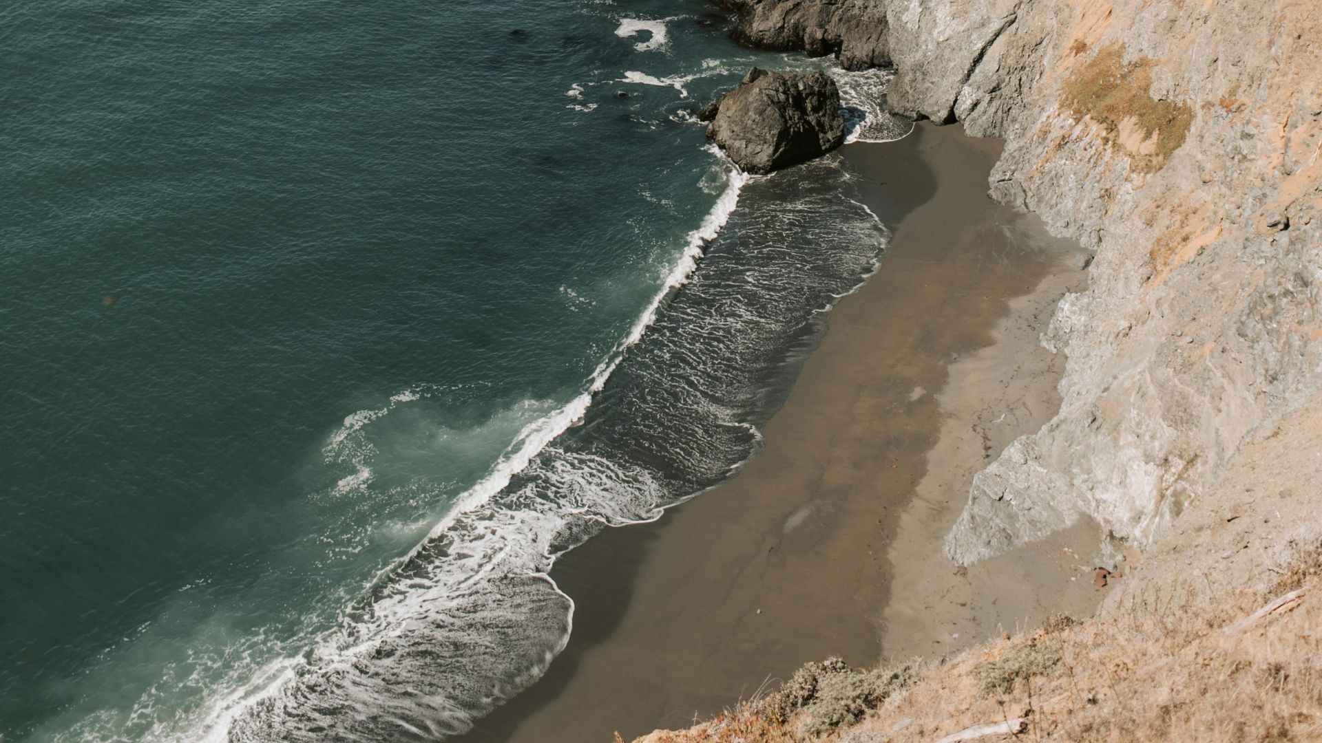 A high-angle view of a secluded beach at the base of steep, rocky cliffs, with waves breaking on the dark sand and the vast ocean stretching into the distance.