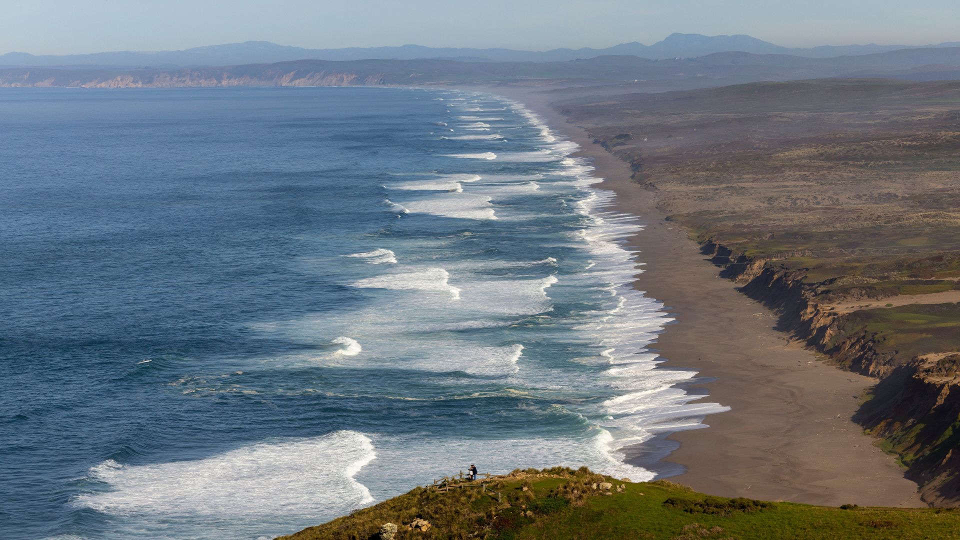 A high-angle view captures the expansive coastline of Point Reyes National Seashore, showcasing a long sandy beach with breaking waves extending into the distance, bordered by a green, grassy cliff in the foreground where a lone figure stands, and hazy mountains in the background.