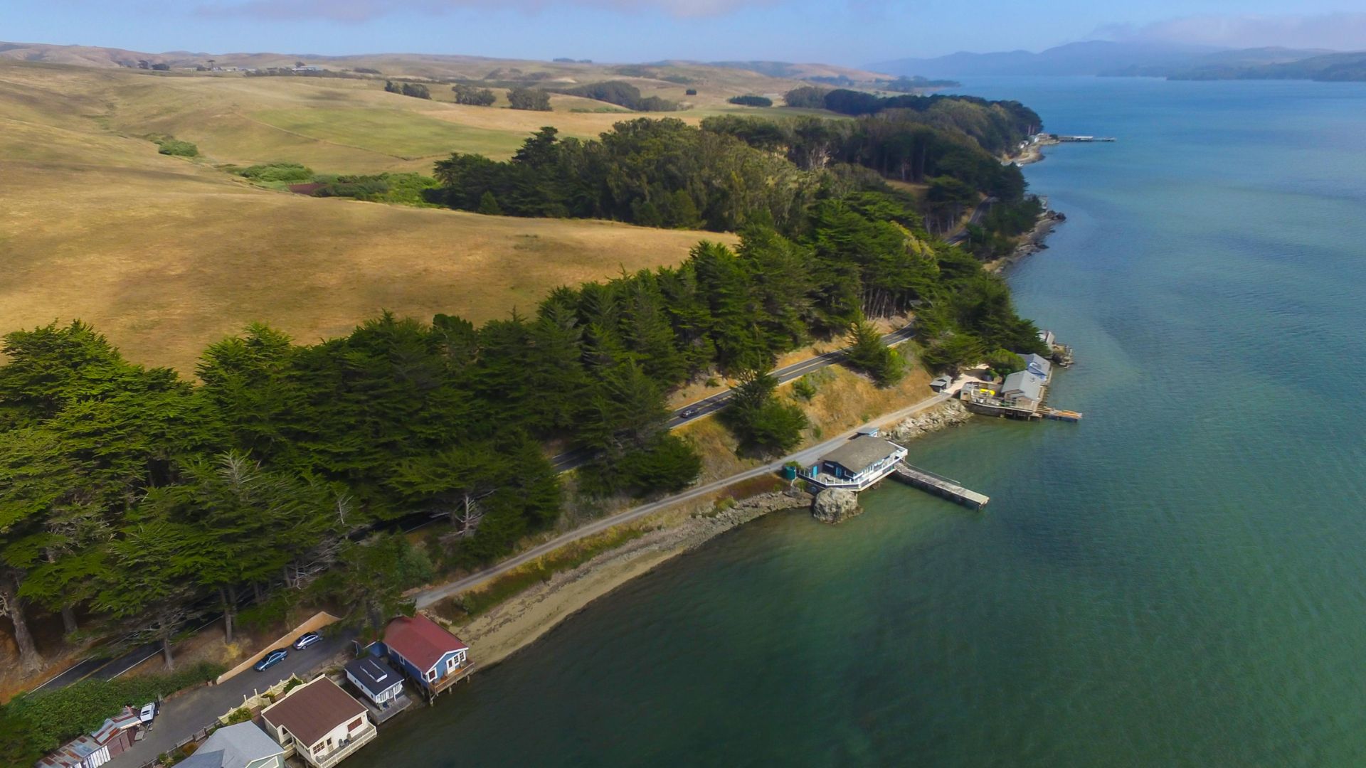 An aerial view of a scenic coastline featuring a narrow strip of land with houses and a road alongside a body of water, backed by rolling hills and forested areas, with a pier and boathouse extending into the water.