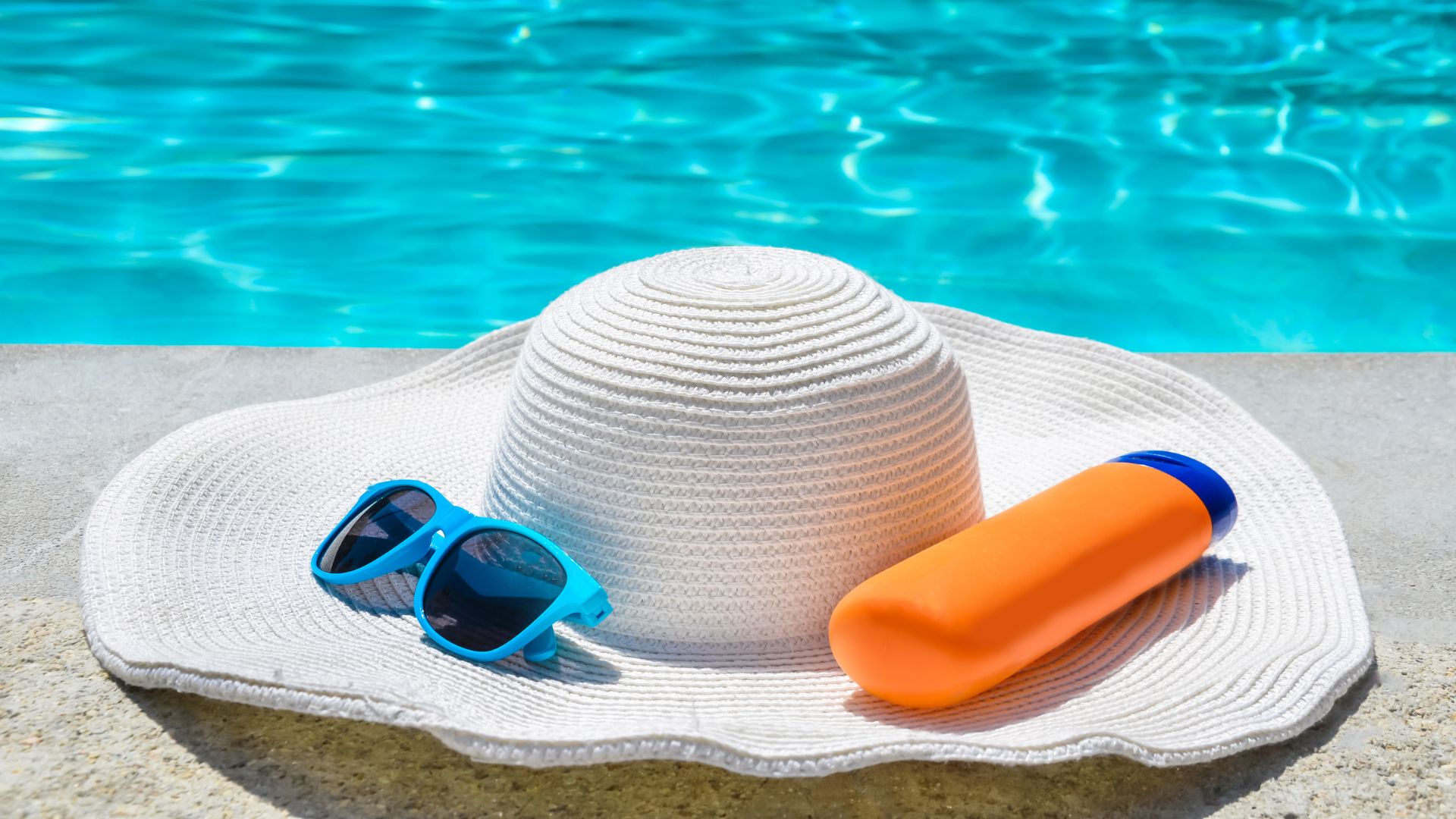 A white sun hat with a pair of blue sunglasses and an orange sunscreen bottle resting on its brim, positioned on the edge of a swimming pool.