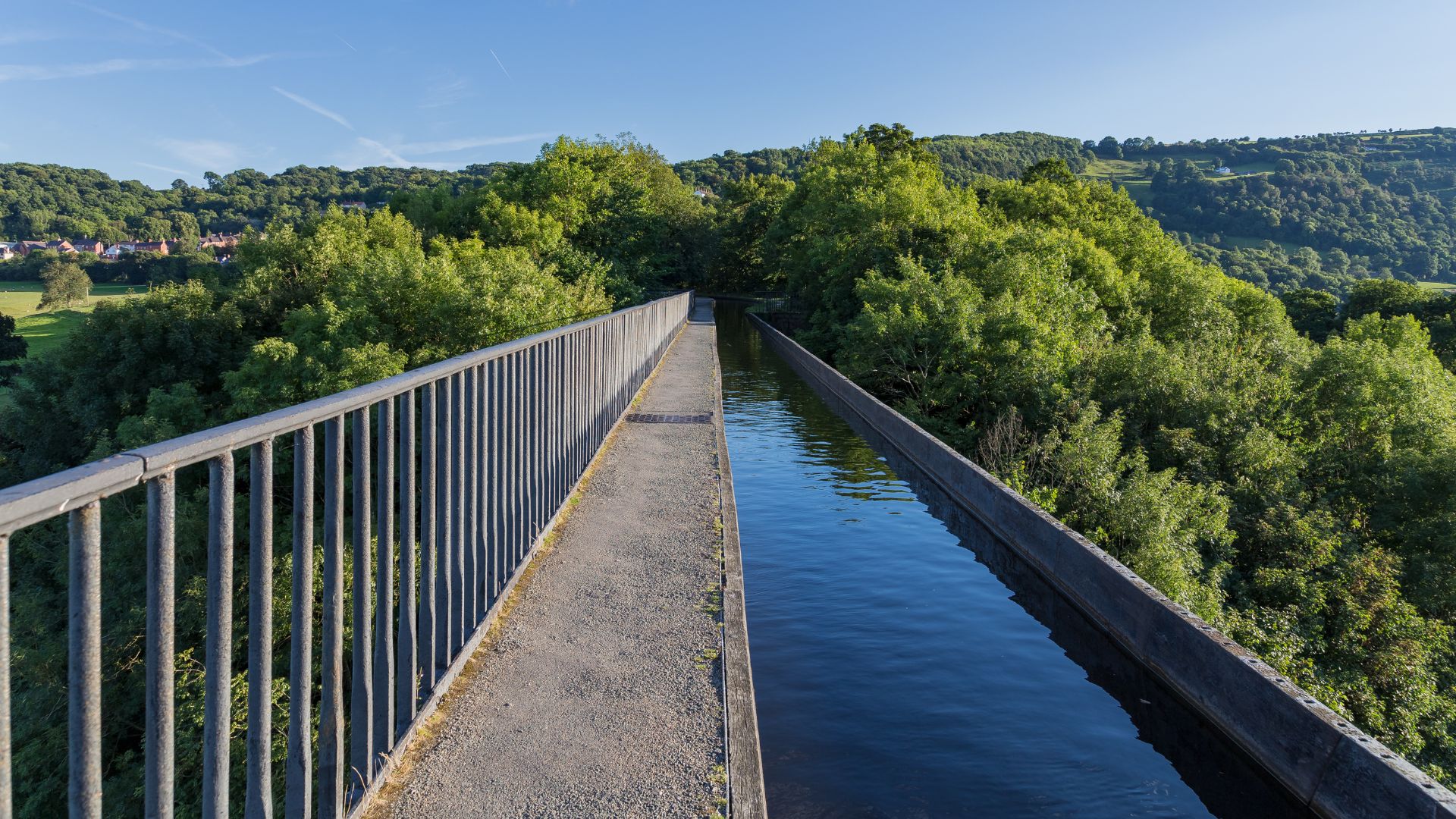 A long, narrow aqueduct carrying a canal of water, with a walkway and railing running alongside it, stretching across a lush, green valley under a clear blue sky.