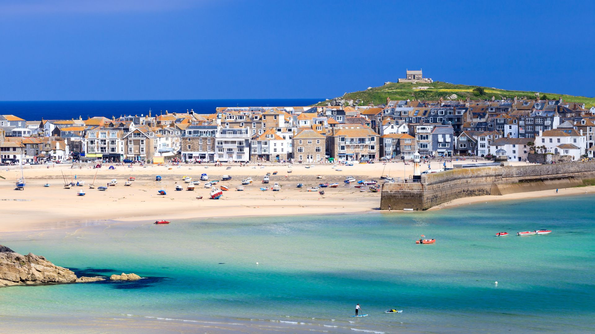 A vibrant aerial view of Porthminster Beach in St Ives, Cornwall, showing turquoise waters, a sandy beach with scattered boats, a stone harbor wall, and a hillside town with numerous buildings, topped by a prominent structure on the distant hill.