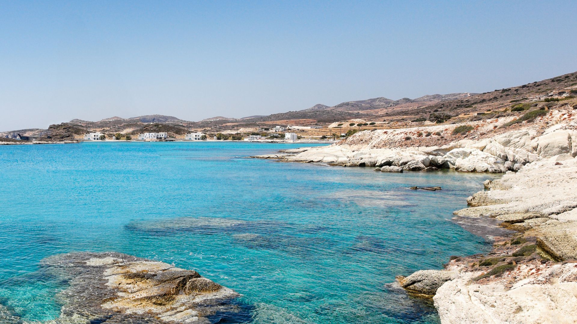 A wide-angle view of Kimolos, Greece, featuring a sandy shoreline meeting clear turquoise waters, with a rocky headland on the left and a barren, hilly landscape rising in the background under a bright blue sky.