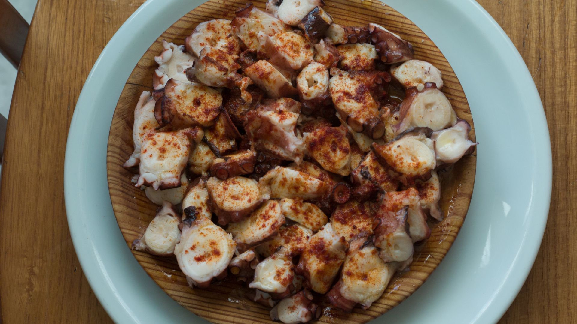 The image displays a close-up overhead shot of a dish featuring chopped octopus tentacles, generously dusted with paprika and possibly drizzled with olive oil, served on a rustic wooden plate set within a light-colored ceramic plate.