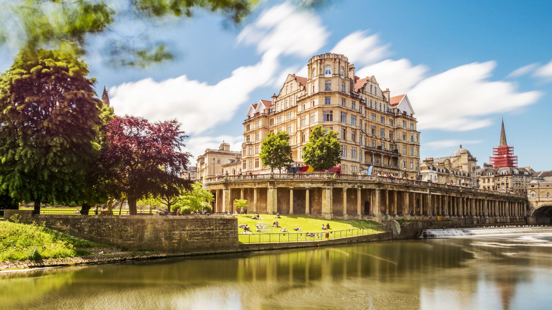 A wide-angle shot of the historic Pulteney Bridge in Bath, England, spanning the River Avon. The bridge features shops built along its full span and is backed by a large, ornate building (the Empire Hotel) with classical architecture and a distant spire visible on the right. Lush green trees and grass line the riverbanks in the foreground, with reflections visible in the calm water. The sky is partly cloudy.