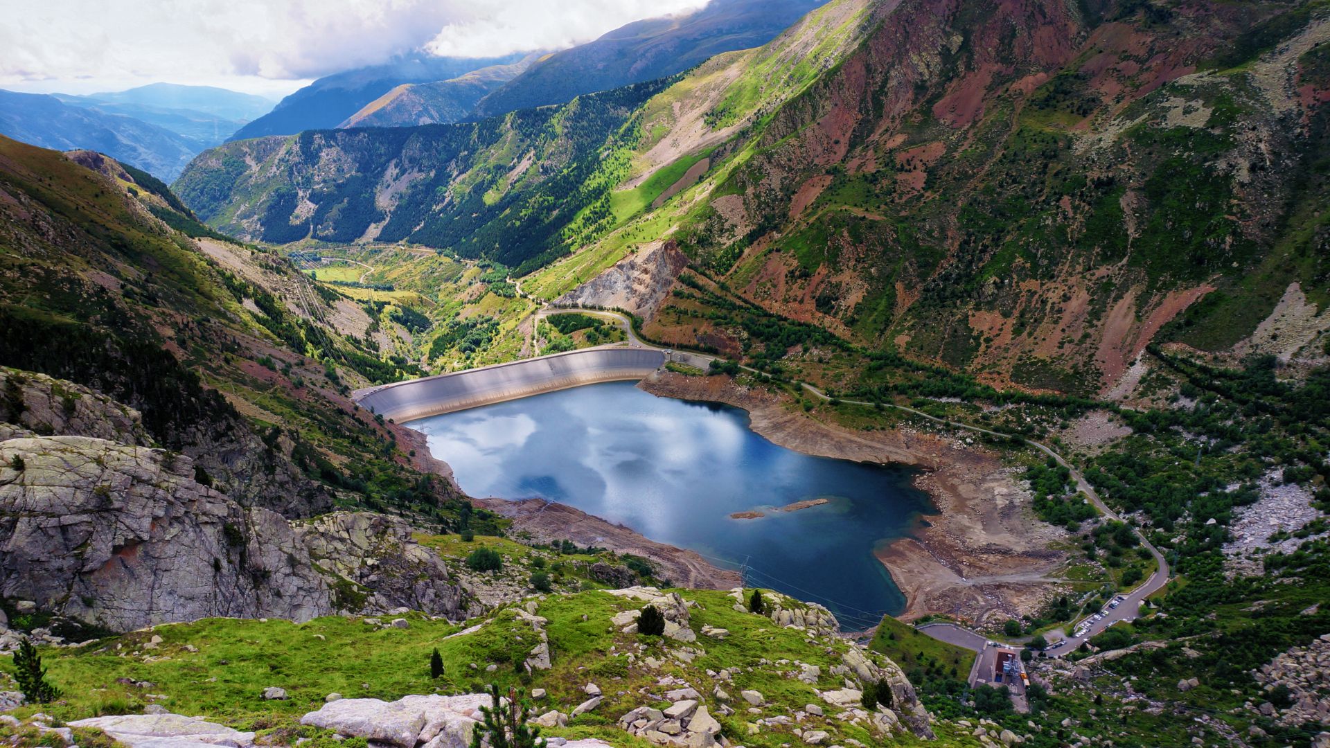 A high-angle shot reveals a deep blue mountain lake or reservoir nestled amidst steep, rugged green and brown mountains under a cloudy sky, with a visible dam structure and a winding road on the right side.