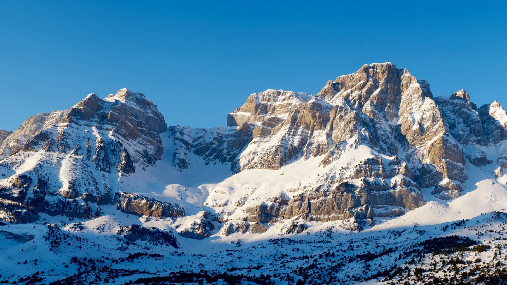 Pyrenees, within Huesca, Aragon, SpainA wide shot of snow-capped mountains under a clear blue sky, with the lower slopes covered in deep snow and the rocky peaks illuminated by sunlight.