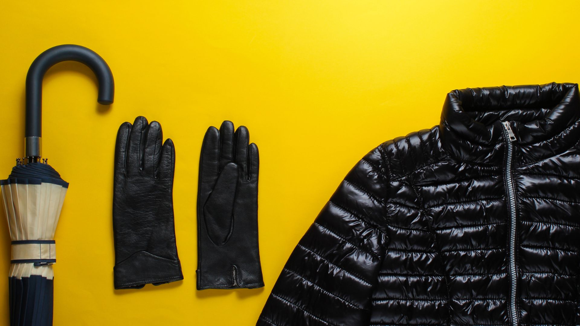 A black umbrella, a pair of black leather gloves, and a black puffer jacket are arranged on a bright yellow background.