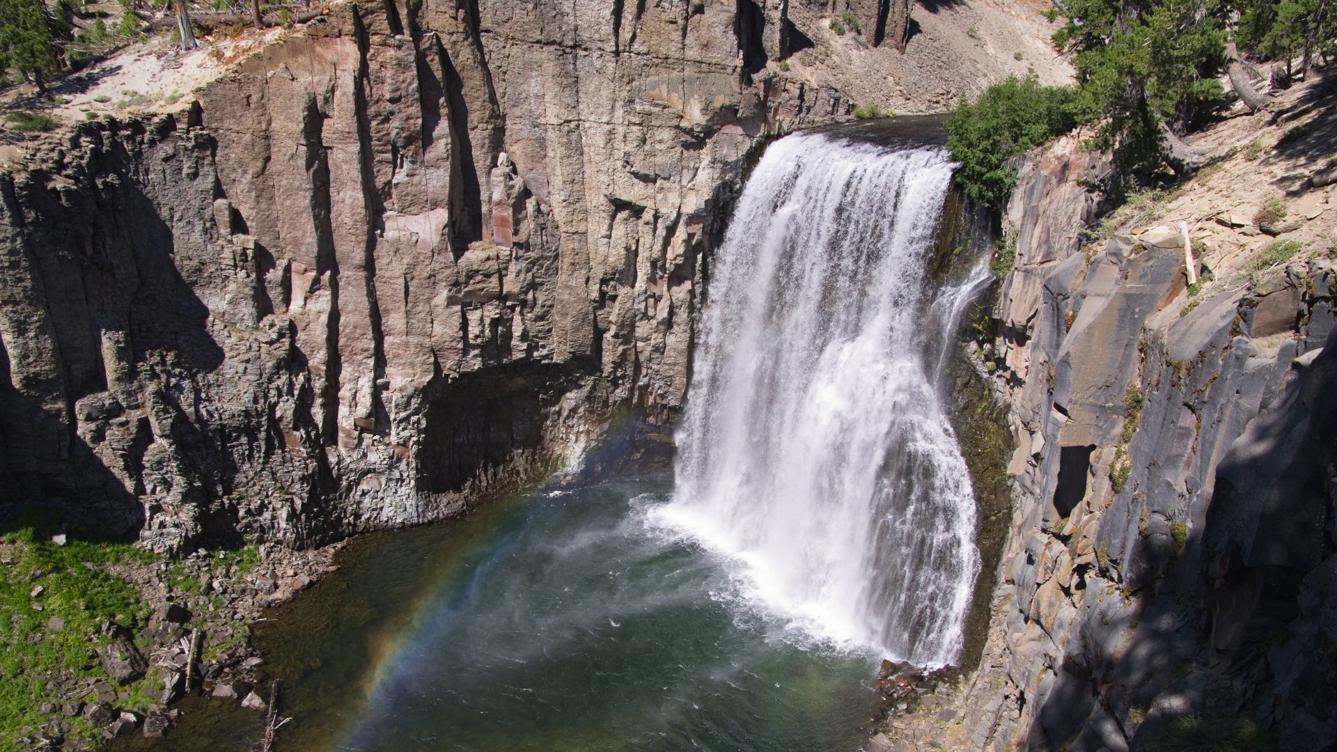 A powerful waterfall cascades down a rocky cliff face into a pool, with a vibrant rainbow visible in the mist at the base of the falls.
