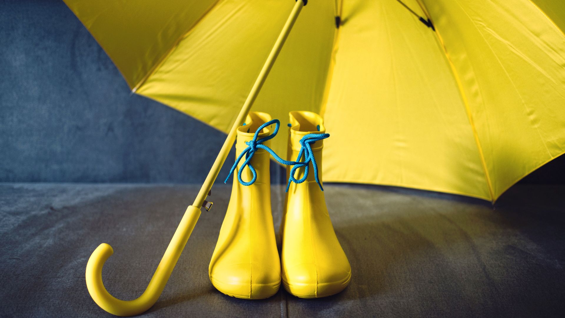A yellow umbrella stands next to a pair of yellow rain boots with blue laces, set against a dark background.