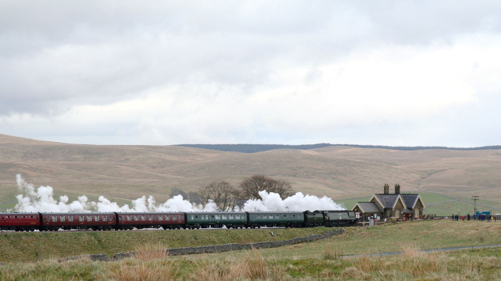 A steam train emitting billowing white smoke pulls into a rural train station with a stone building, surrounded by rolling green hills under a cloudy sky.