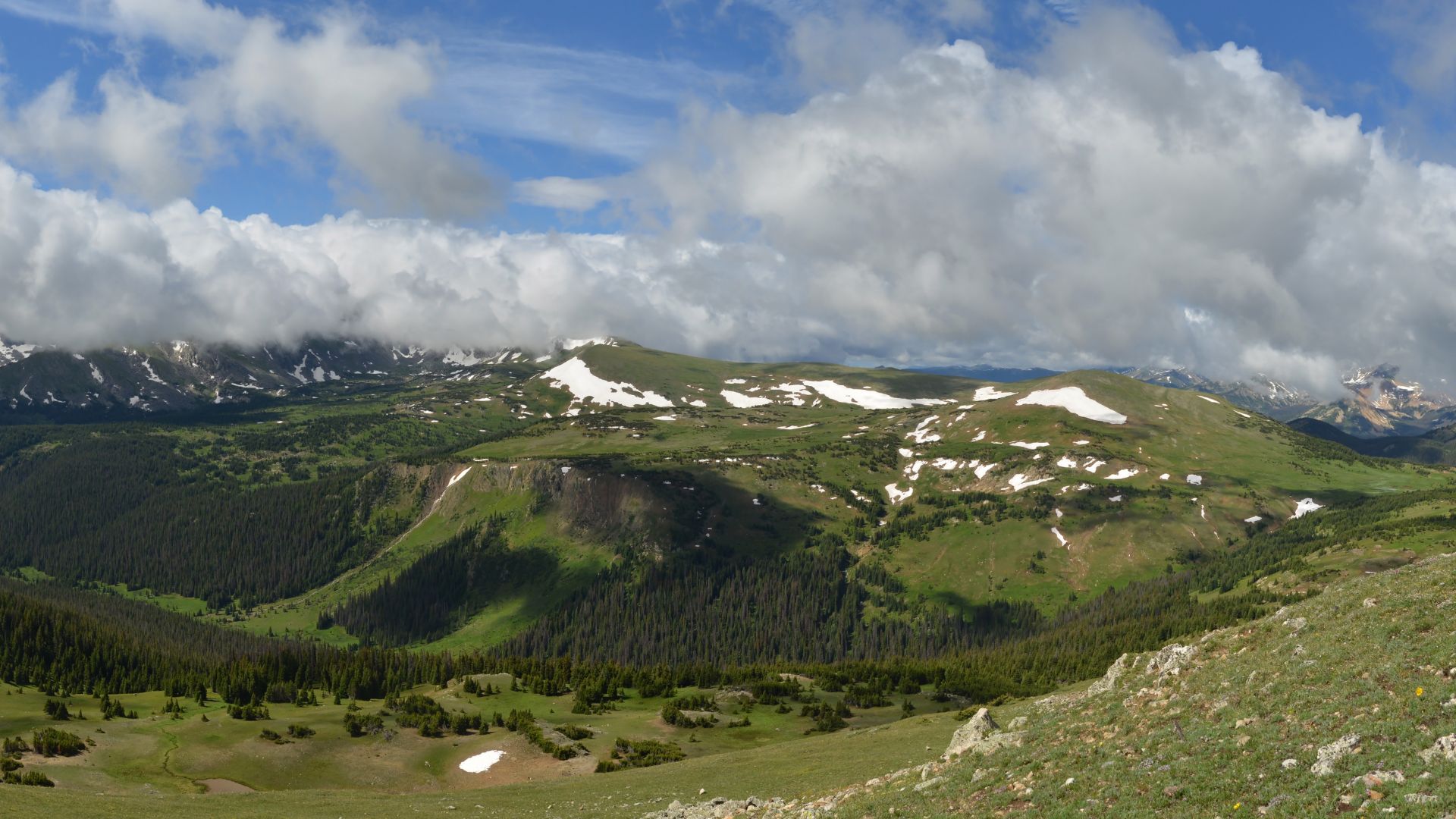 A panoramic view of a vast alpine meadow landscape in the Rocky Mountains, featuring green rolling hills, patches of snow, scattered evergreen trees, and a dramatic sky filled with white clouds.
