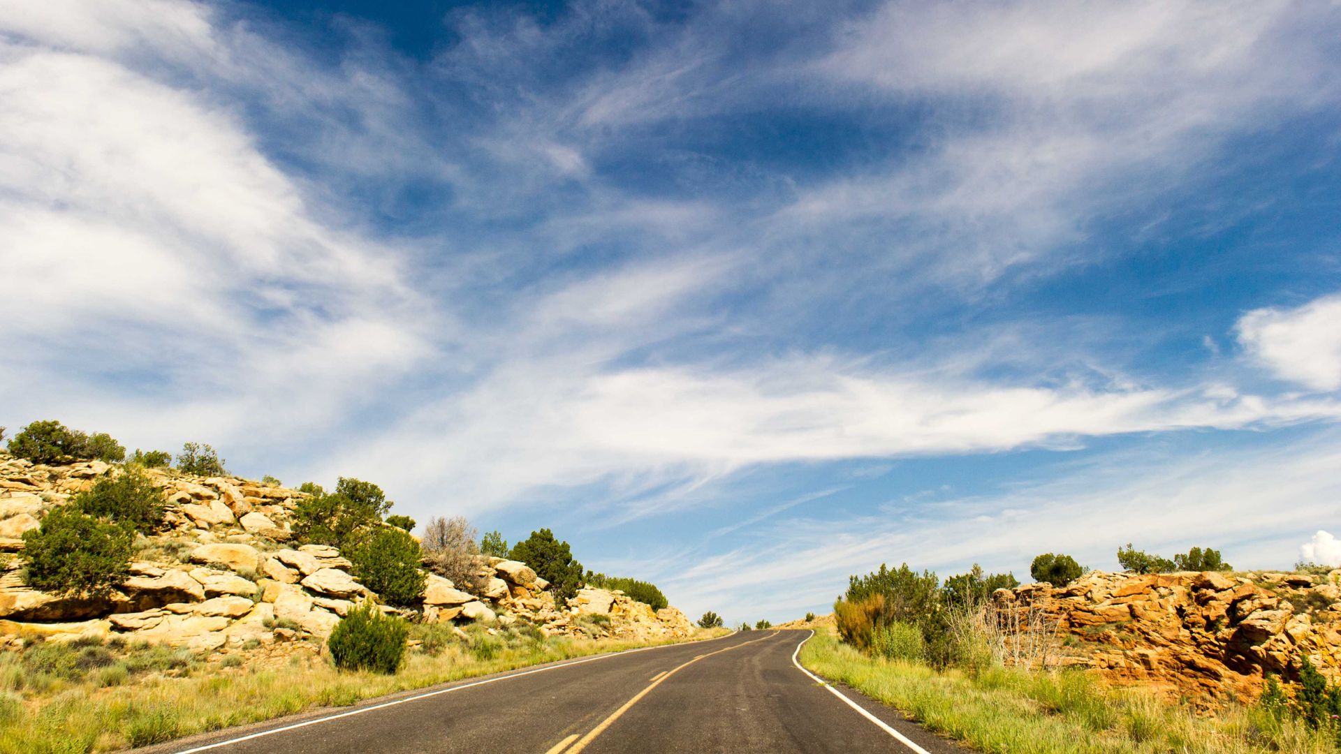 A long, winding road stretches through a desert landscape with rocky hills and sparse green vegetation under a wide blue sky with wispy clouds.
