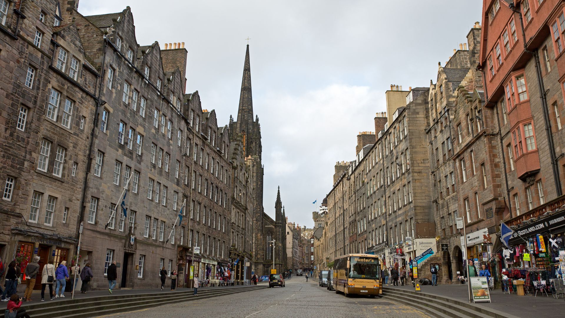 A view down a bustling, historic street lined with tall, old stone buildings, featuring a prominent church spire in the distance and a double-decker bus on the cobbled road.